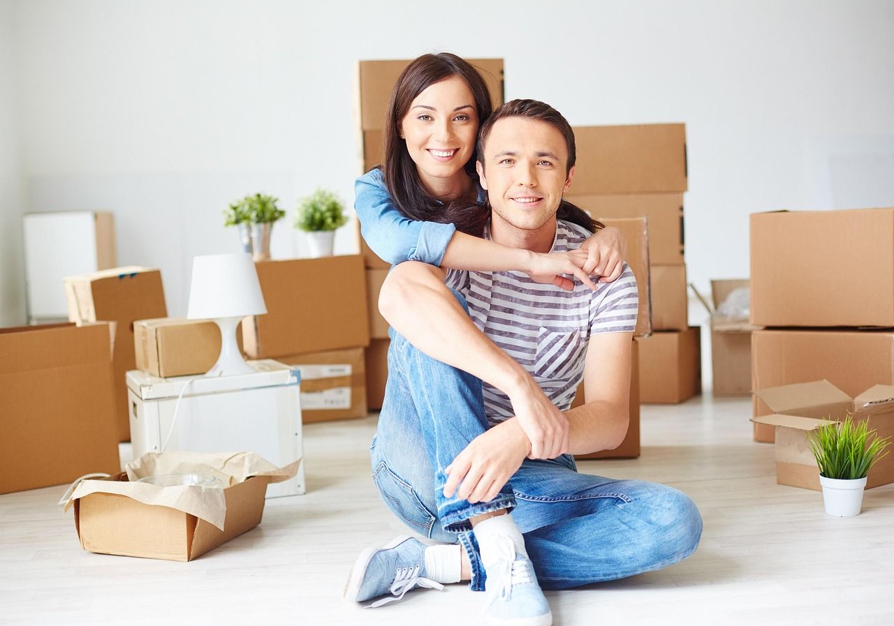 A couple hugging and smiling while sitting down, surrounded by house moving boxes.