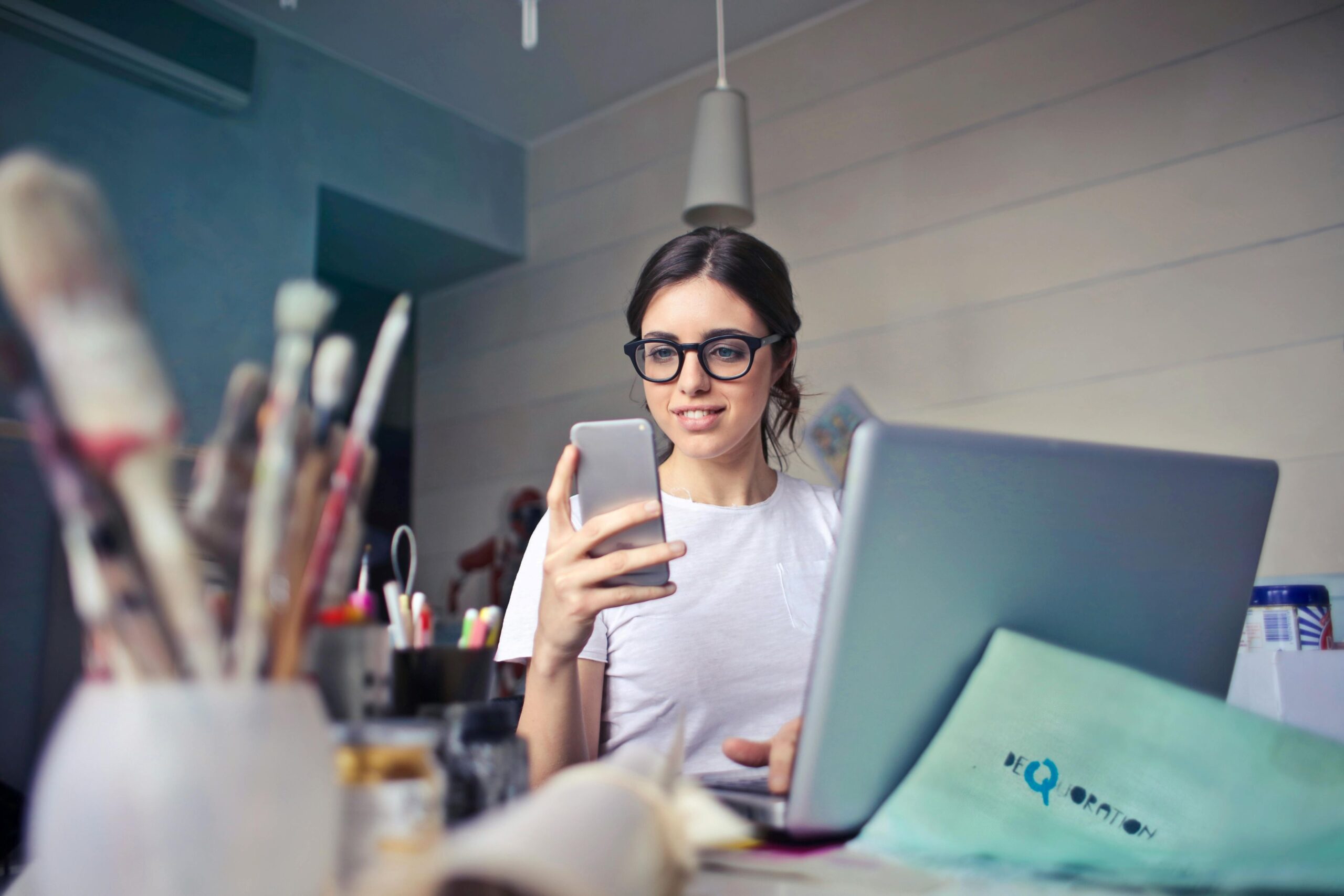 A female wearing glasses is checking her phone, with laptop in front, surrounded by art materials.